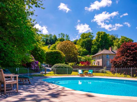 An outdoor swimming pool with lounge chairs and a fenced garden with trees and a house in the background at Haytor Cottage in Cornworthy