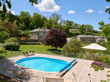 An outdoor swimming pool with lounge chairs and an umbrella table in a garden with trees and houses in the background at Yennadon Cottage in Cornworthy