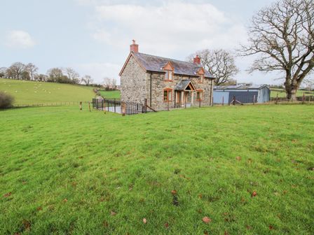 A stone house with orange window frames on a grassy field near a metal shed and trees at Ty Gwyn in Llanfair Caereinion