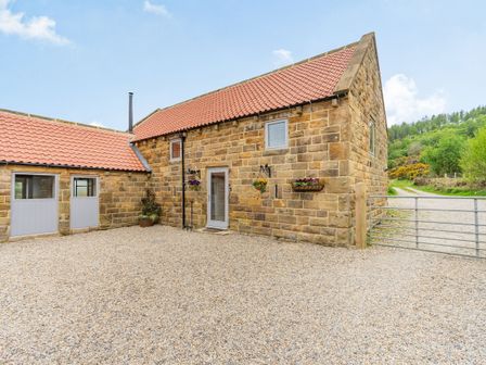 A stone building with a tiled roof and a gravel driveway with flower baskets on the wall at Meadowsweet in Goathland
