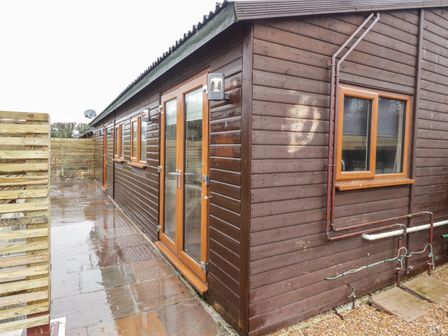 An exterior view of a brown wooden building with multiple windows and doors on a wet stone pavement at Sparrows Den Lodge in Winchelsea Beach