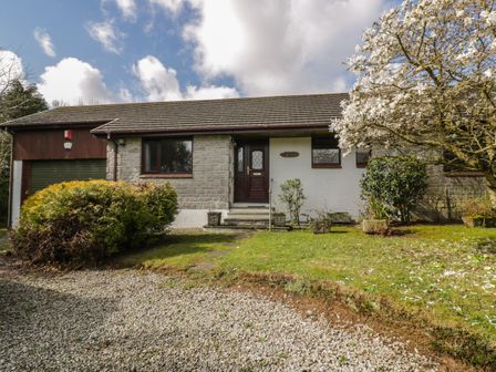 A front view of a single-story house with a gravel driveway and a garden with bushes and a flowering tree at Three Jays in Lanreath