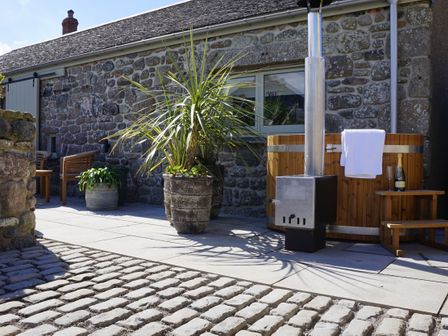 An outdoor patio with a cobblestone and paved floor a wooden hot tub with a towel and champagne bottle a potted plant and wooden chairs by a stone house at The Wink Cape Cornwall