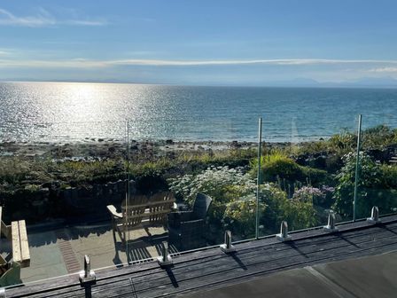 A patio with wooden benches and glass railing overlooking the sea and rocky shore at Red Ridge in Llandanwg near Harlech