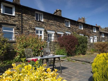 A stone cottage exterior with a patio table and two chairs surrounded by bushes and flowers at Dormouse Cottage in Coniston