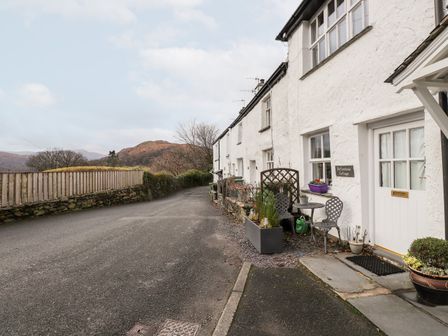 A row of white cottages along a narrow road with outdoor chairs and potted plants at Buttonhole Cottage in Ambleside
