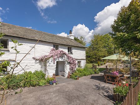 Exterior of a white cottage with flowers around entrance and outdoor seating under an umbrella at Smithy Cottage At Lindeth in Bowness