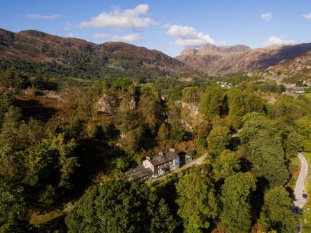 A house surrounded by trees and mountains under a partly cloudy sky at The Stable At Oakbank in Elterwater