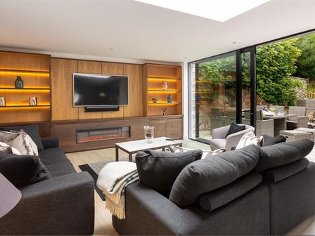 A living room with dark sofas a wall-mounted tv illuminated shelves and large glass doors opening to an outdoor seating area at Underhowe in Grasmere