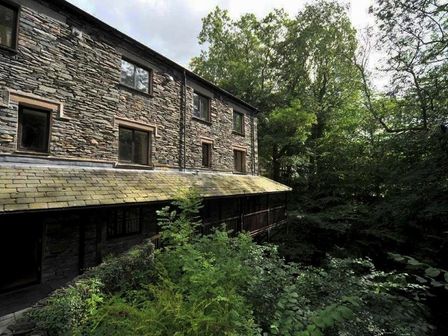 A stone building with multiple windows surrounded by dense green trees and plants at River Falls View in Ambleside