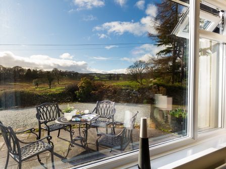 An outdoor patio with a metal table and four chairs set with a plant and cups overlooking a green landscape with trees and a blue sky at Honister Cottage in Ambleside
