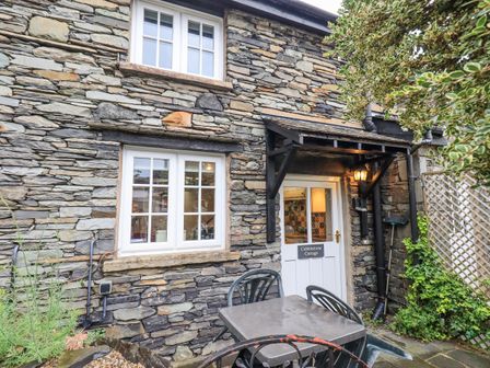 The exterior of a stone cottage with two windows a white door with a canopy and a small patio area with a table and chairs at Cobblestone Cottage in Ambleside