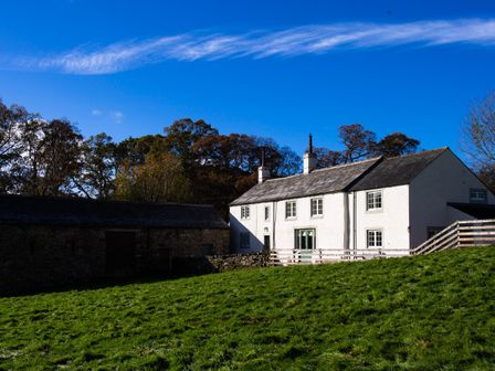 A white farmhouse with chimneys next to a stone barn and green grass under a blue sky at Horrockwood Farm in Watermillock
