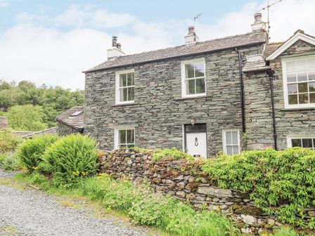 A stone house with white framed windows and a white door behind a stone wall with plants at The Smithy in Keswick