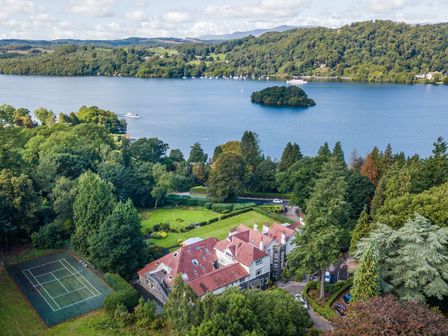 A large house with a tennis court surrounded by trees near a lake with an island and boats at Skyfall in Bowness