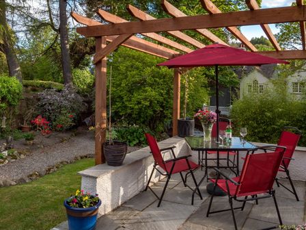 An outdoor patio with a glass table and four red chairs under a wooden pergola at Artists Hideaway in Keswick