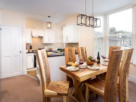 A dining area with wooden table and chairs set with wine glasses and plates next to a kitchen with white cabinets and tiled backsplash at Lingmell House in Bowness