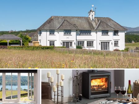A white two-story house with a pitched roof behind a wooden fence and a field of tall grass at Hawkrigg Farm Hawkshead