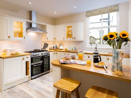 A kitchen with white cabinets wooden countertops a stove a window and a wooden breakfast bar with stools at Brandelhow House Keswick