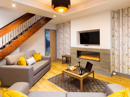 A living room with two grey sofas with yellow pillows a wooden coffee table with a laptop and beer bottles a wall-mounted fireplace and TV a wooden staircase and a door with wallpaper at Sunnybank Cottage in Keswick