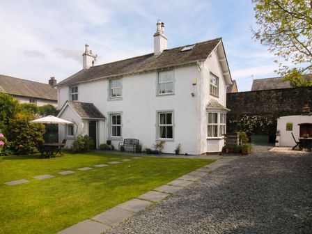 A white two-story house with chimneys and multiple windows with a gravel driveway and green lawn with a table and umbrella at Crow Cottage in Keswick