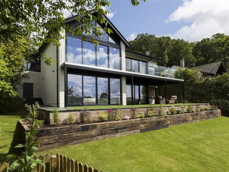 A modern house with large glass windows and a balcony overlooking a garden with a stone retaining wall at Jaspers Retreat in Bowness