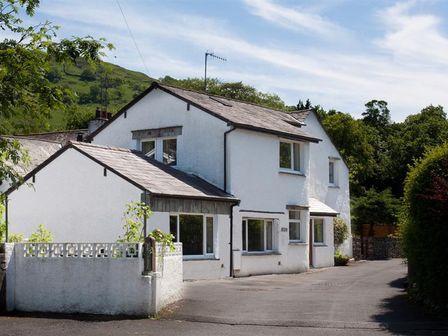 A white two-story house with multiple windows and a small wall in front surrounded by trees at Oaklea in Ambleside