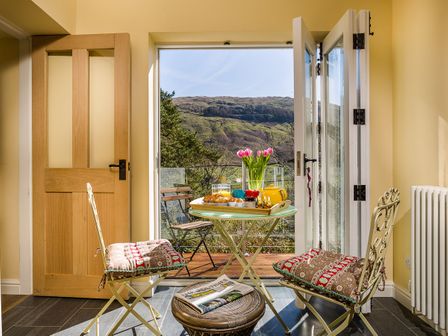 A dining area with a small round table set for breakfast and two chairs with cushions at South Stonethwaite Cottage in Troutbeck