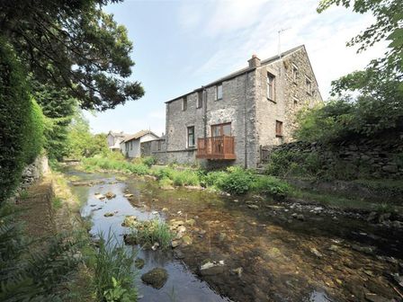 A stone house beside a shallow stream with greenery along the banks at A River Runs By in Staveley