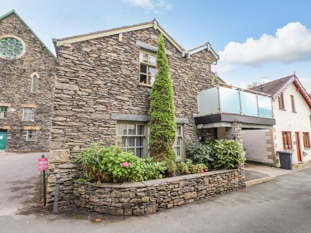 A stone building with a small garden and a balcony with glass railings at Workshop Cottage in Bowness