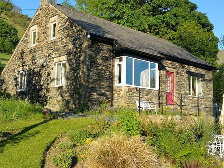A stone house with white framed windows and a red door surrounded by grass and plants at Cherry Garth in Hartsop