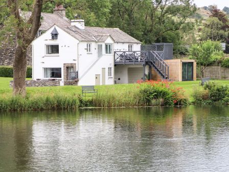 A white house with a wooden deck and stairs next to a body of water with green grass and benches at Riverside Hideaway in Newby Bridge