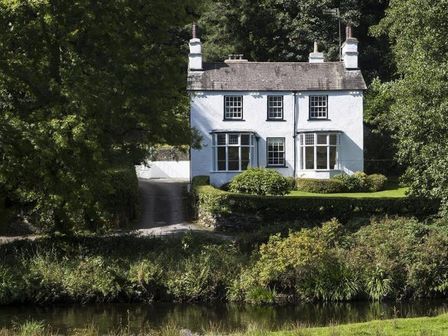A white two story house with chimneys beside a river surrounded by trees at Loughrigg Cottage in Ambleside