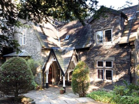 A stone house with a pitched roof and arched doorway surrounded by trees and bushes at Priory Lodge in Windermere