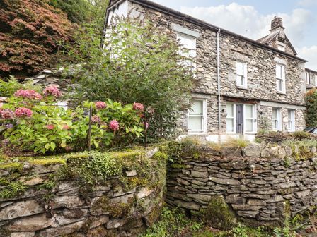 A stone house with white window frames and a front door behind a mossy stone wall with plants and pink flowers at The Mews in Ambleside