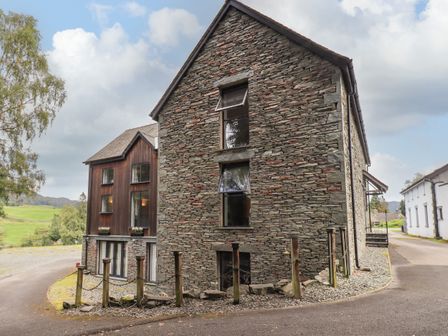 A stone and wooden building beside a curved road with trees and grassy hills in the background at Le Chalet in Elterwater