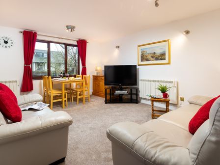 A living room with beige sofas and red cushions a wooden dining table with chairs a television and a window with red curtains at High Lake in Bowness