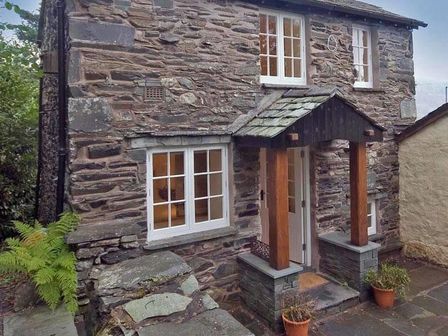 The stone exterior of a cottage with white framed windows a small porch with wooden pillars and potted plants at Ivy Cottage in Ambleside