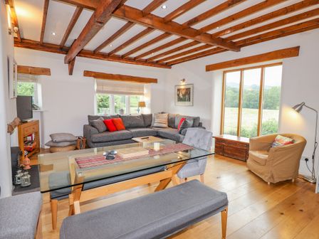A living room with a gray sectional sofa red cushions a glass dining table wooden beams on ceiling and large windows at Glen View Cottage in Grasmere