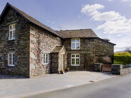 A stone house with white framed windows and a small porch on a paved driveway at Tilly's Cottage in Grasmere