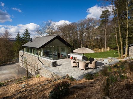 A stone house with large windows and a patio with outdoor furniture and umbrella at Tall Trees in Bowness