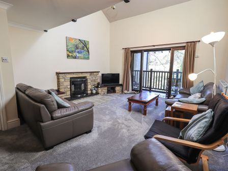 A living room with leather sofas a wooden coffee table a stone fireplace and sliding glass doors leading to a balcony at The Spinney in Ambleside