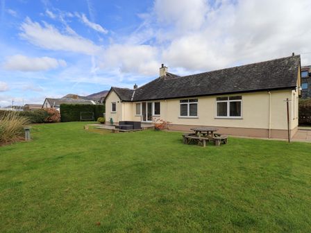 A beige single-story house with a dark tiled roof and lawn with wooden picnic table in the garden at Laal Field House in Keswick