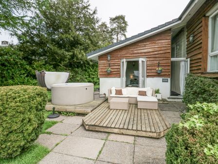 An outdoor patio with a wooden deck sofa hot tub and surrounding bushes at Lake Lodge in Bowness-On-Windermere