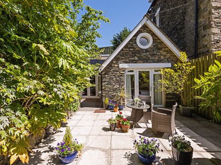A stone house with a patio area containing a table and chairs and potted plants at Wheatlands Cottage in Windermere