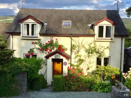 A two-story cottage with a stone roof surrounded by bushes and flowers at Garden Cottage At Coniston