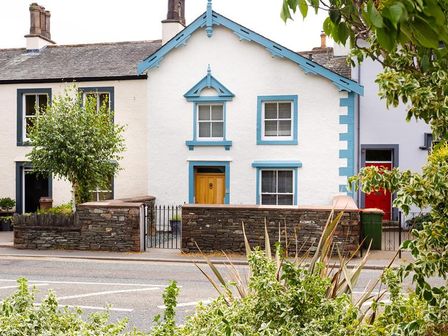 A white two-story house with blue trim and wooden door behind a stone wall with a metal gate at Pippin Cottage in Keswick