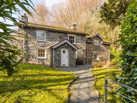 A stone cottage with a white door and gravel path in front surrounded by grass and trees at Thimble Hall in Grasmere