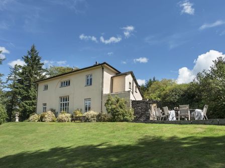 A large house with white walls surrounded by bushes and trees with an outdoor table and chairs on a stone patio at Tent Lodge in Coniston