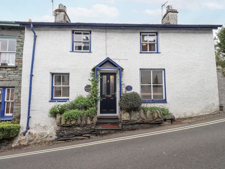 A white cottage with blue window frames and a black door on a sloped street at Springwell Cottage in Ambleside